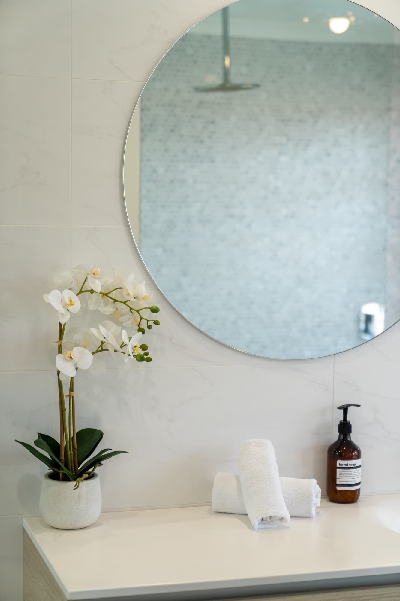 A bathroom with a round mirror, a potted orchid, a hand soap bottle, and two towels on a white countertop.