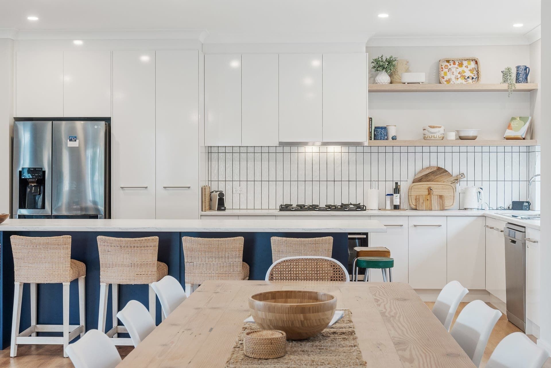 A modern kitchen with white cabinets, a stainless steel refrigerator, a built-in stove, and a dining area with a wooden table and wicker chairs.