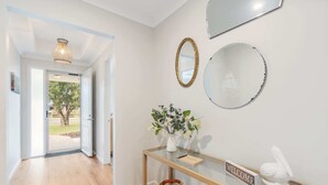A hallway with a wooden floor, a glass console table with a 'WELCOME' sign, a vase with flowers, and a mirror. - Shepherds Delight-Disabled/Access Friendly (Goolwa South)