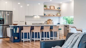 A modern kitchen with white cabinets, a stainless steel refrigerator, a bar area with wicker stools, and a view of greenery outside. - Shepherds Delight-Disabled/Access Friendly (Goolwa South)