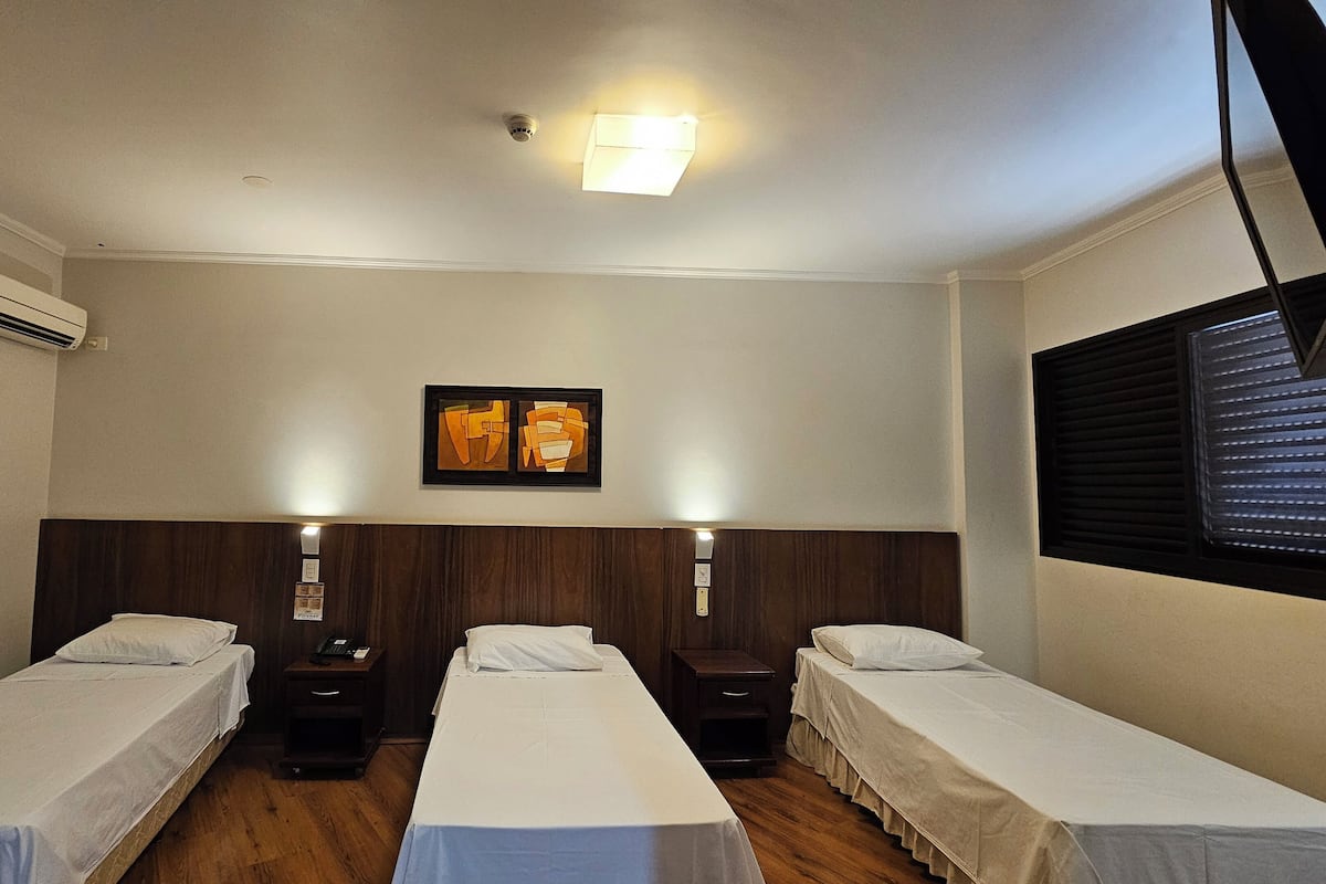 Een hotelkamer met drie enkele bedden, een houten hoofdbord, een aan het plafond bevestigde airconditioner, een lijst met een abstract schilderij en een raam met jaloezieën.