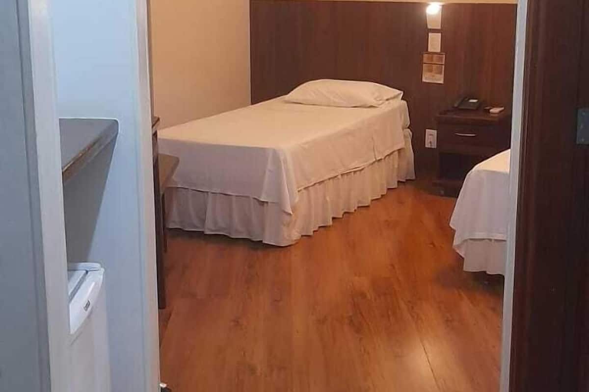Een smalle gang die uitkomt in een slaapkamer met een houten vloer, een bed met een witte dekbedovertrek, een nachtkastje en een wandlamp.