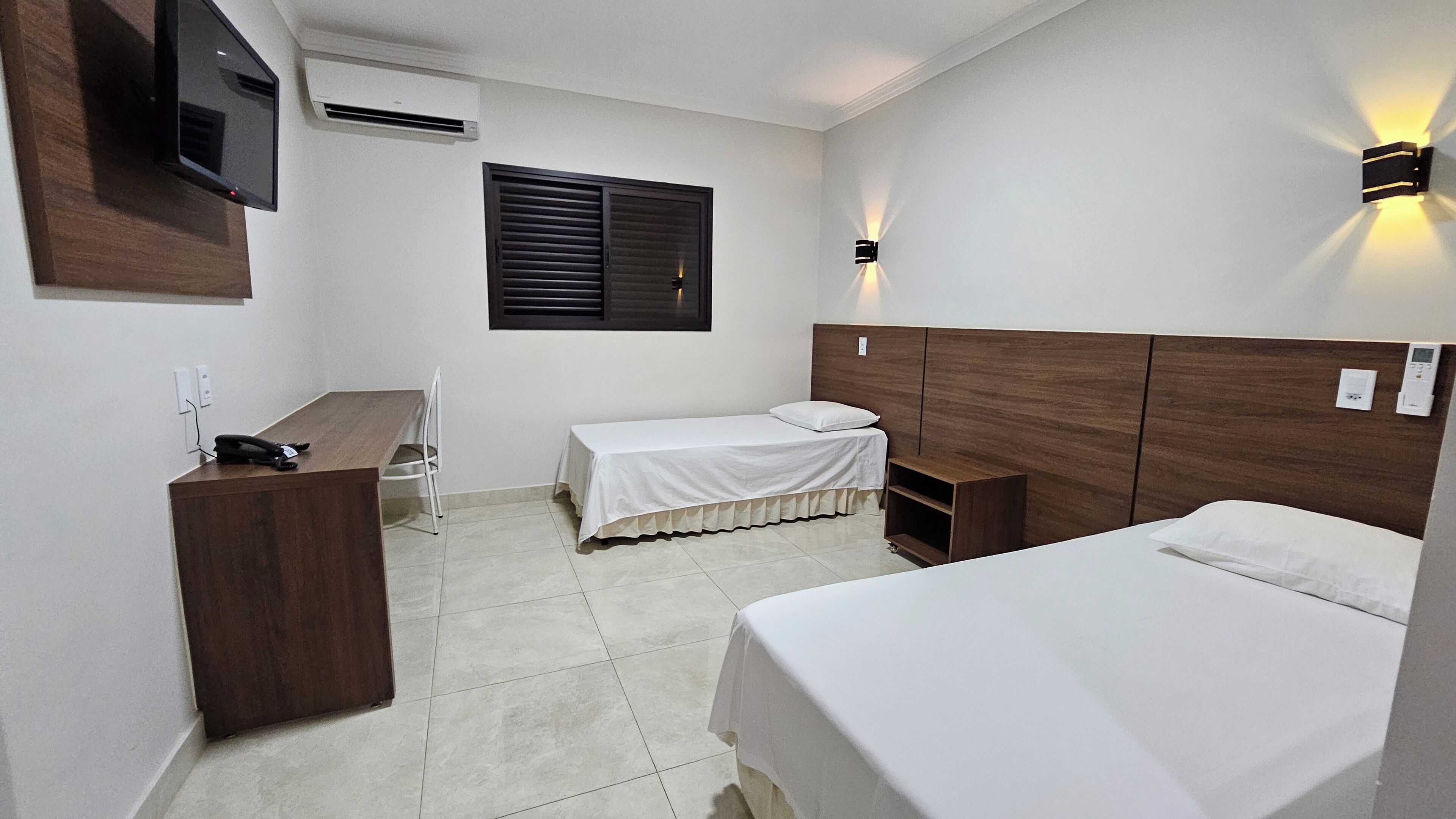 Quarto de hotel com cama, escrivaninha, cadeira, televisão, janela com persianas e luminária fixada na parede.