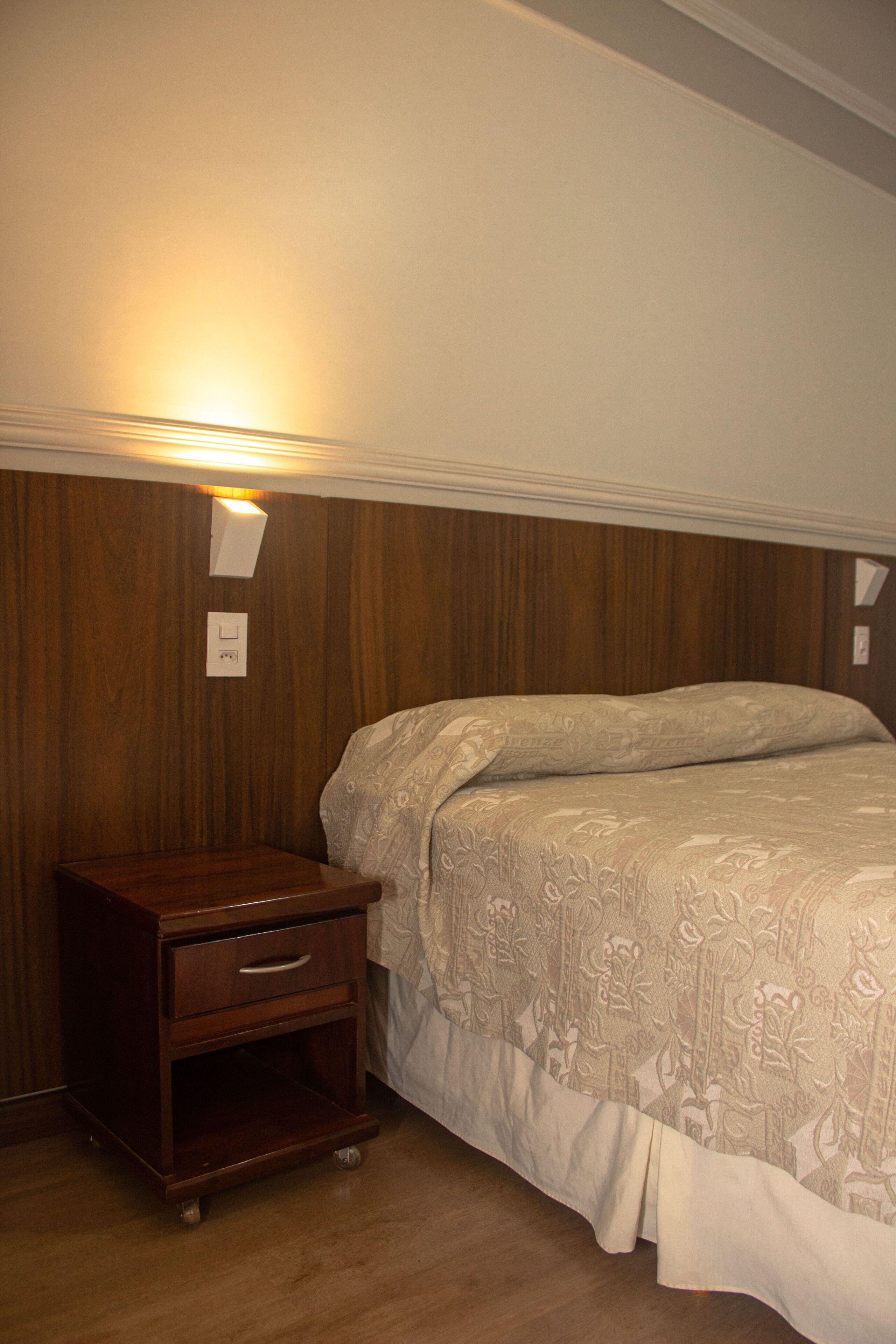 A bedroom with a wooden nightstand, a bed with a patterned bedspread, and a wall-mounted light fixture.
