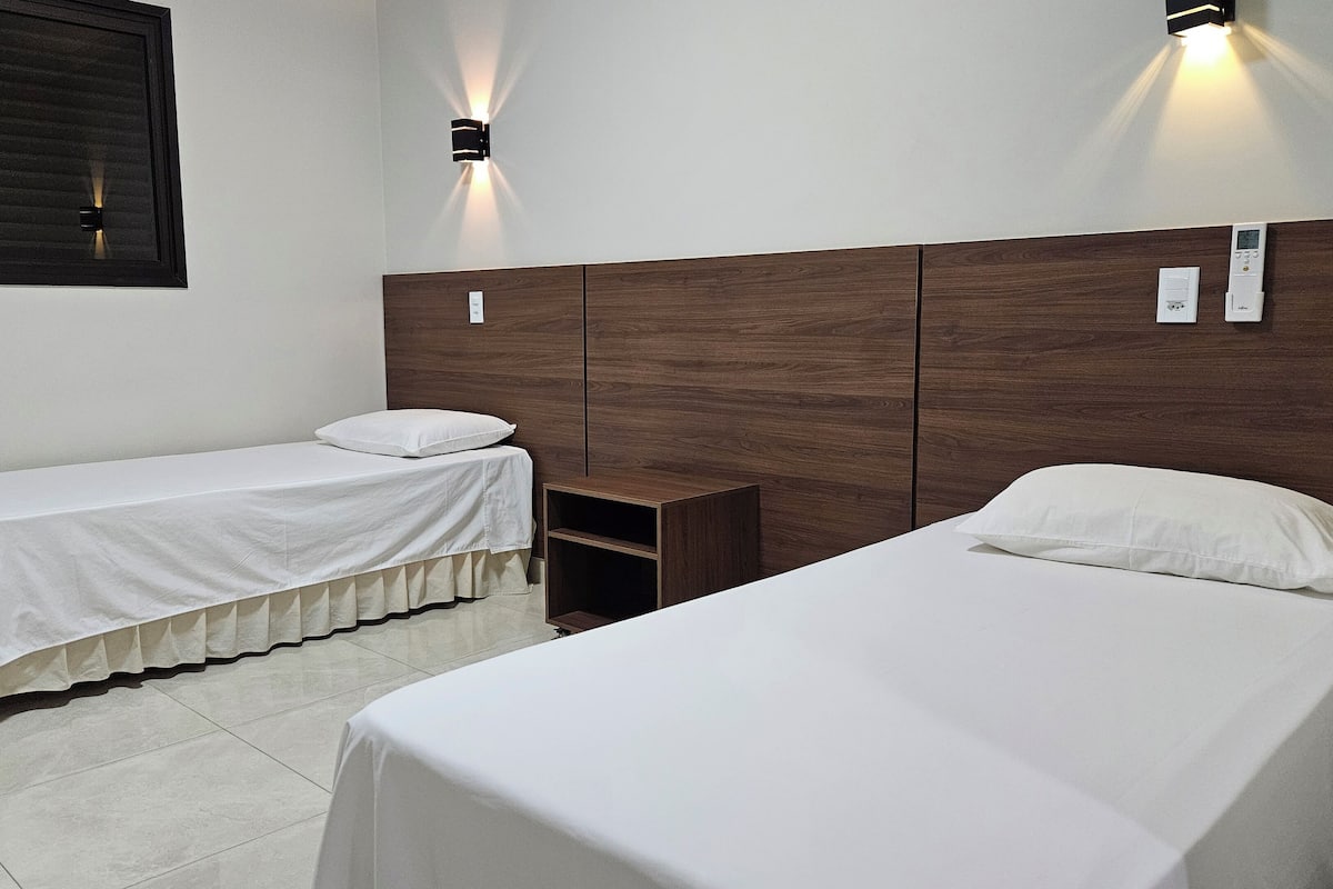 Een hotelkamer met twee eenpersoonsbedden, een houten hoofdbord, een klein nachtkastje van hout en een wandlamp.