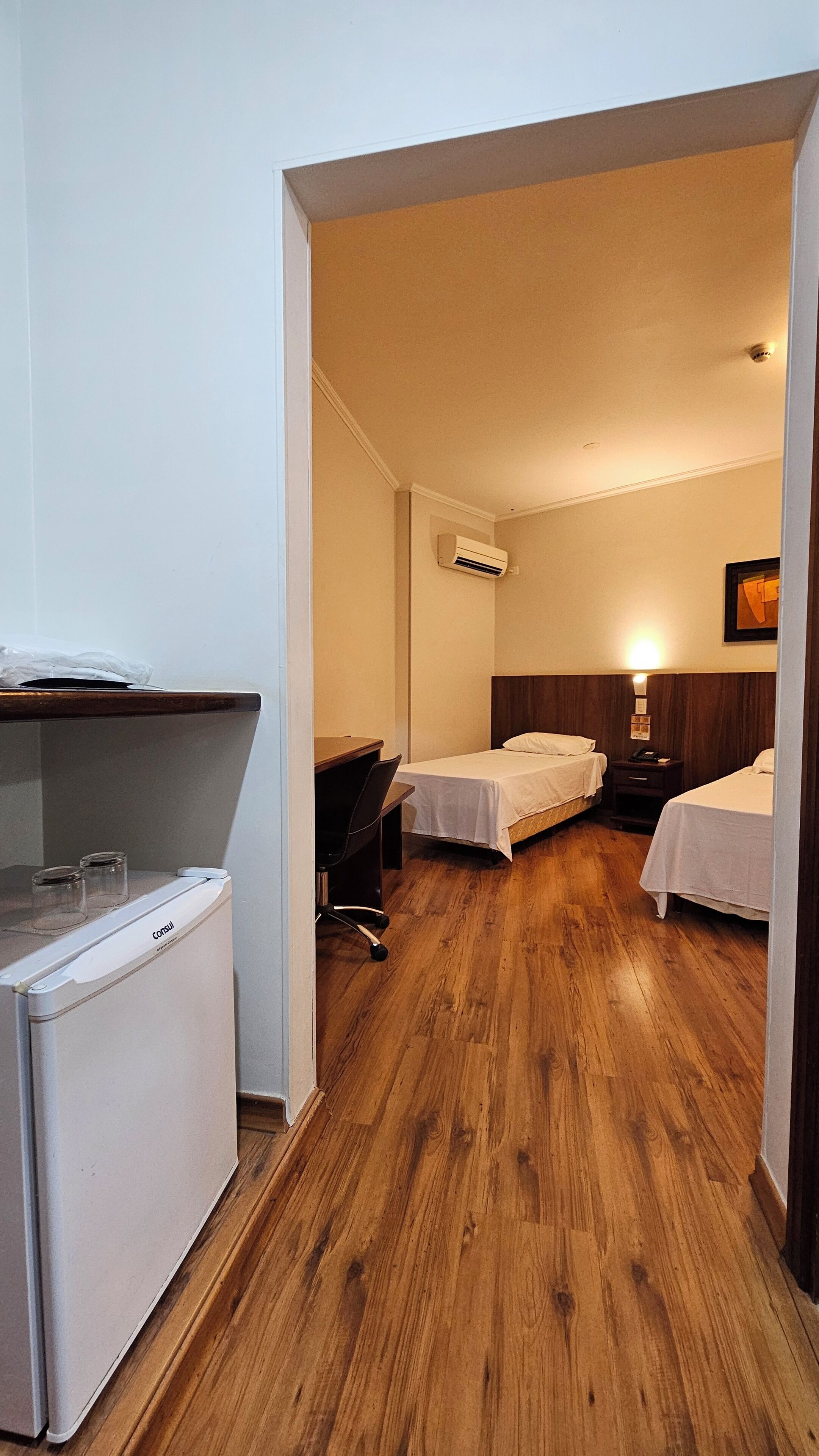 Camera d'albergo con due letti singoli, una scrivania con sedia, un piccolo frigorifero e un condizionatore d'aria.