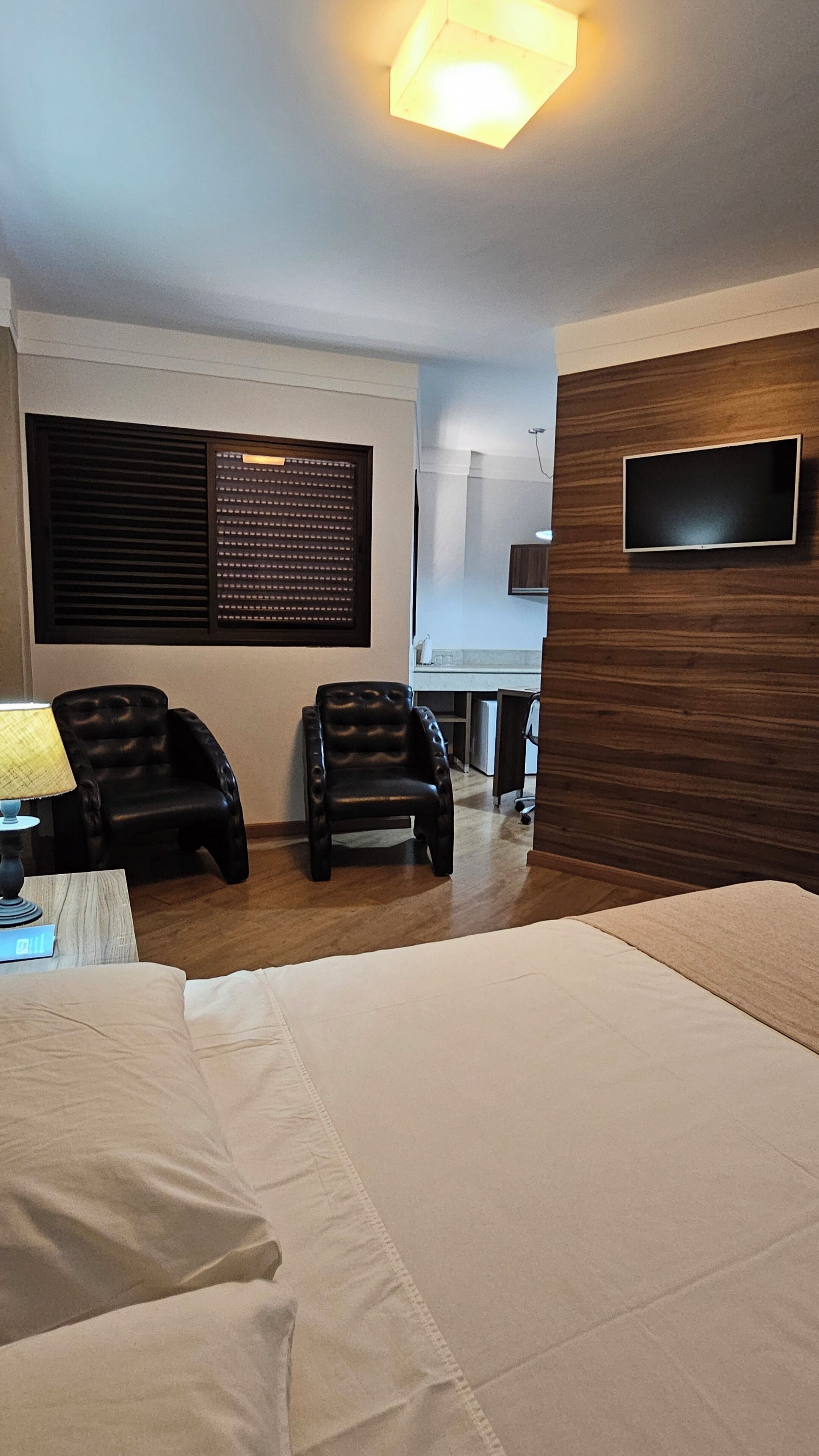 A hotel room with a bed, two leather chairs, a TV mounted on the wall, a window with blinds, and a desk with a lamp.