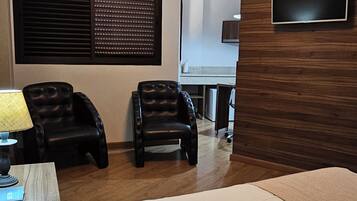 A hotel room with a bed, two leather chairs, a TV mounted on the wall, a window with blinds, and a desk with a lamp.