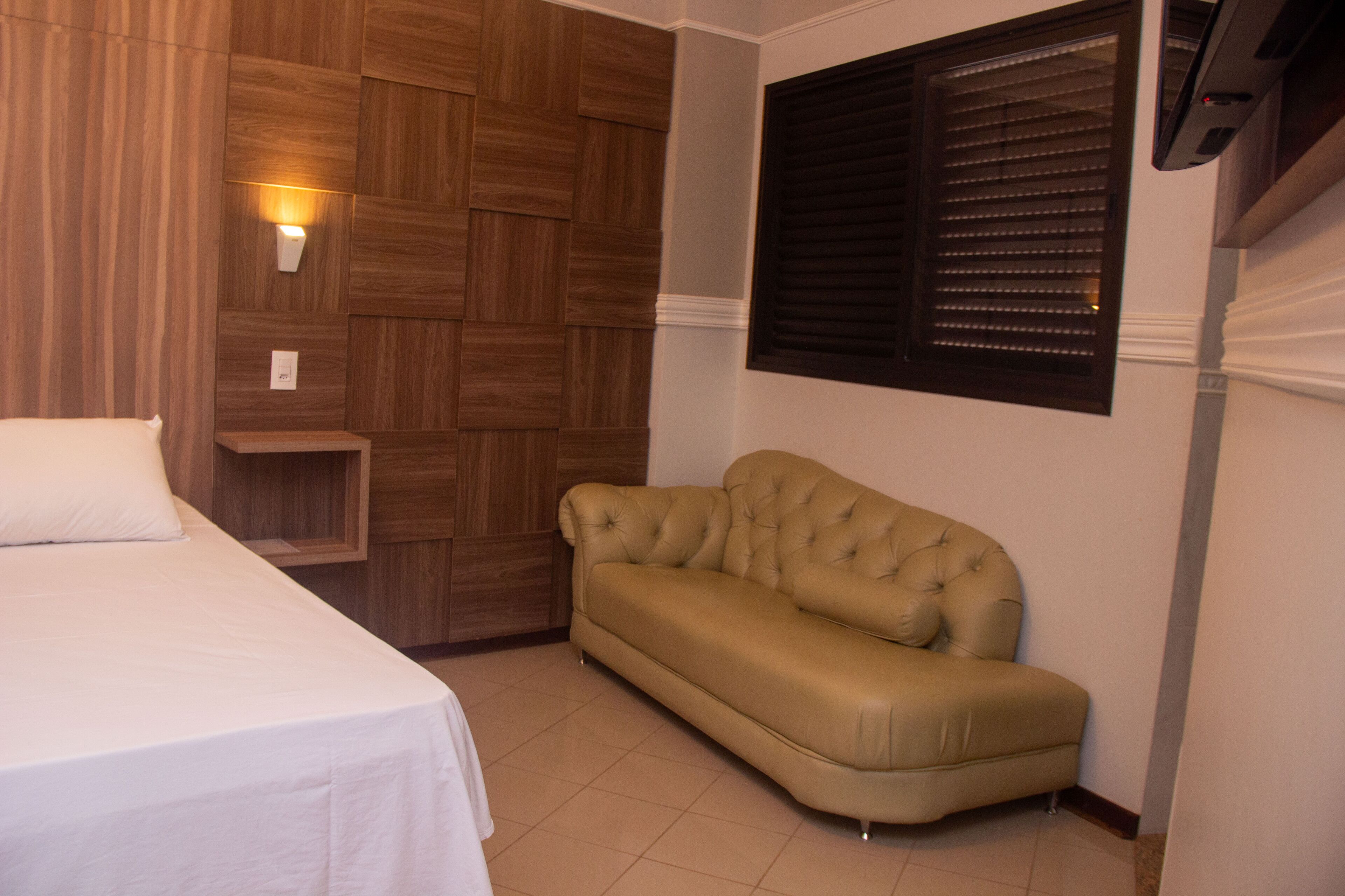 A bedroom with a bed, a beige sofa, wooden paneling, a window with blinds, and a wall-mounted air conditioning unit.