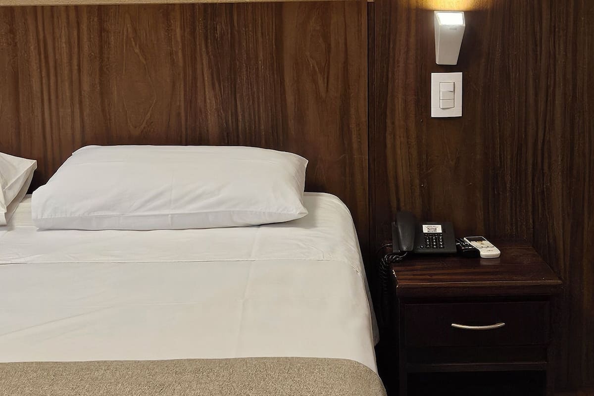 Een hotelkamer met een bed, nachtkastje, lamp en een schilderij van bomen aan de muur.
