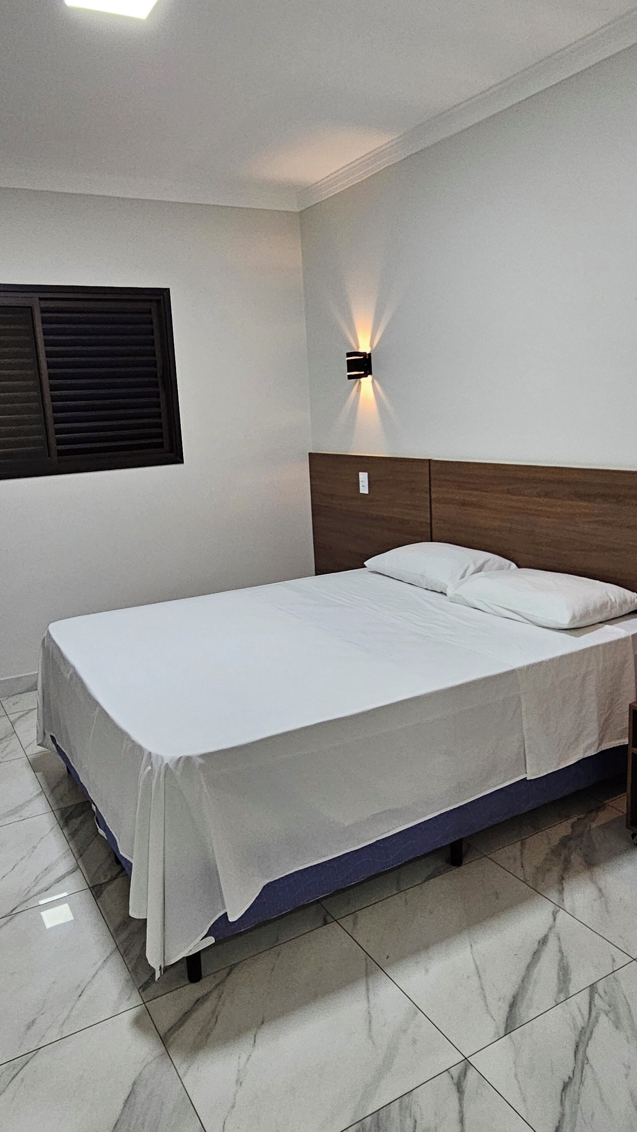A neatly made bed in a room with a tiled floor and a wall-mounted light fixture.