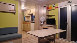 A compact room with a table, two chairs, a sofa, a TV, a fan, and a door marked with an emergency exit sign. - Sunset Milfontes (Odemira)
