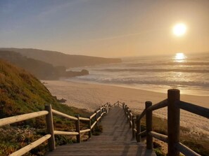Una escalera de madera que lleva a una playa, con baranda de madera, y ofrece vistas al océano y al atardecer.