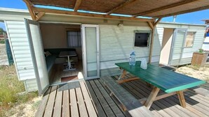 A mobile home with a covered outdoor area, a picnic table, and a sliding door leading to a small room with a bed and a window. - Sunset Milfontes (Odemira)