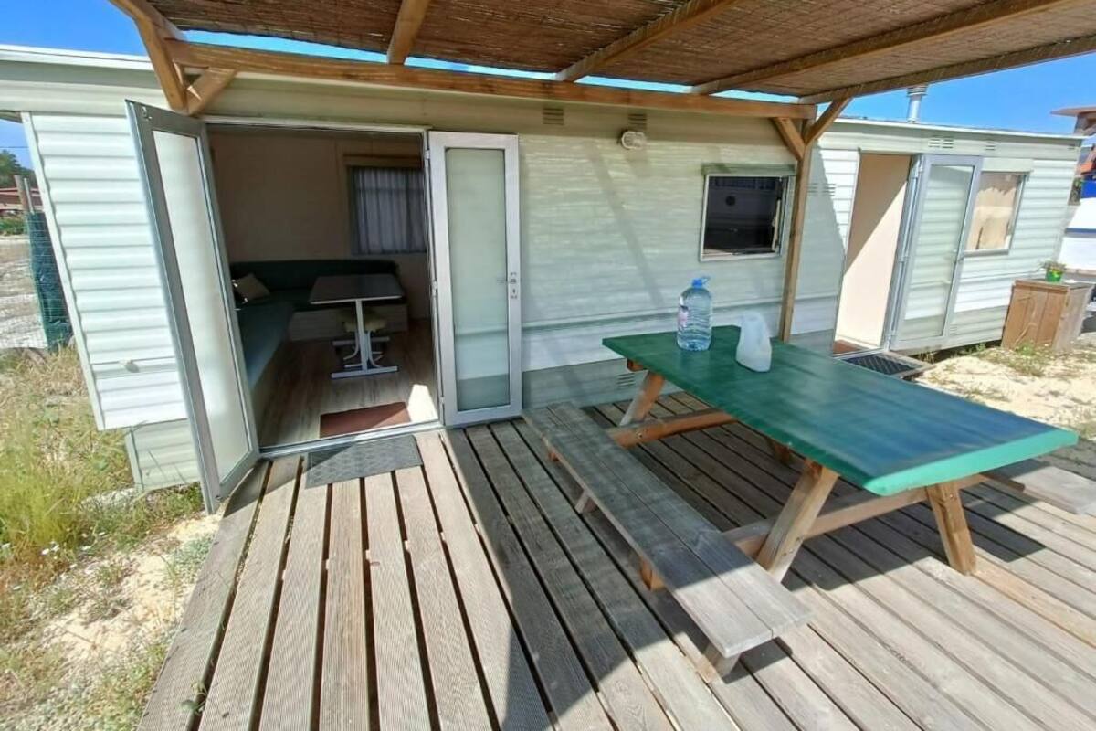 A mobile home with a covered outdoor area, a picnic table, and a sliding door leading to a small room with a bed and a window.