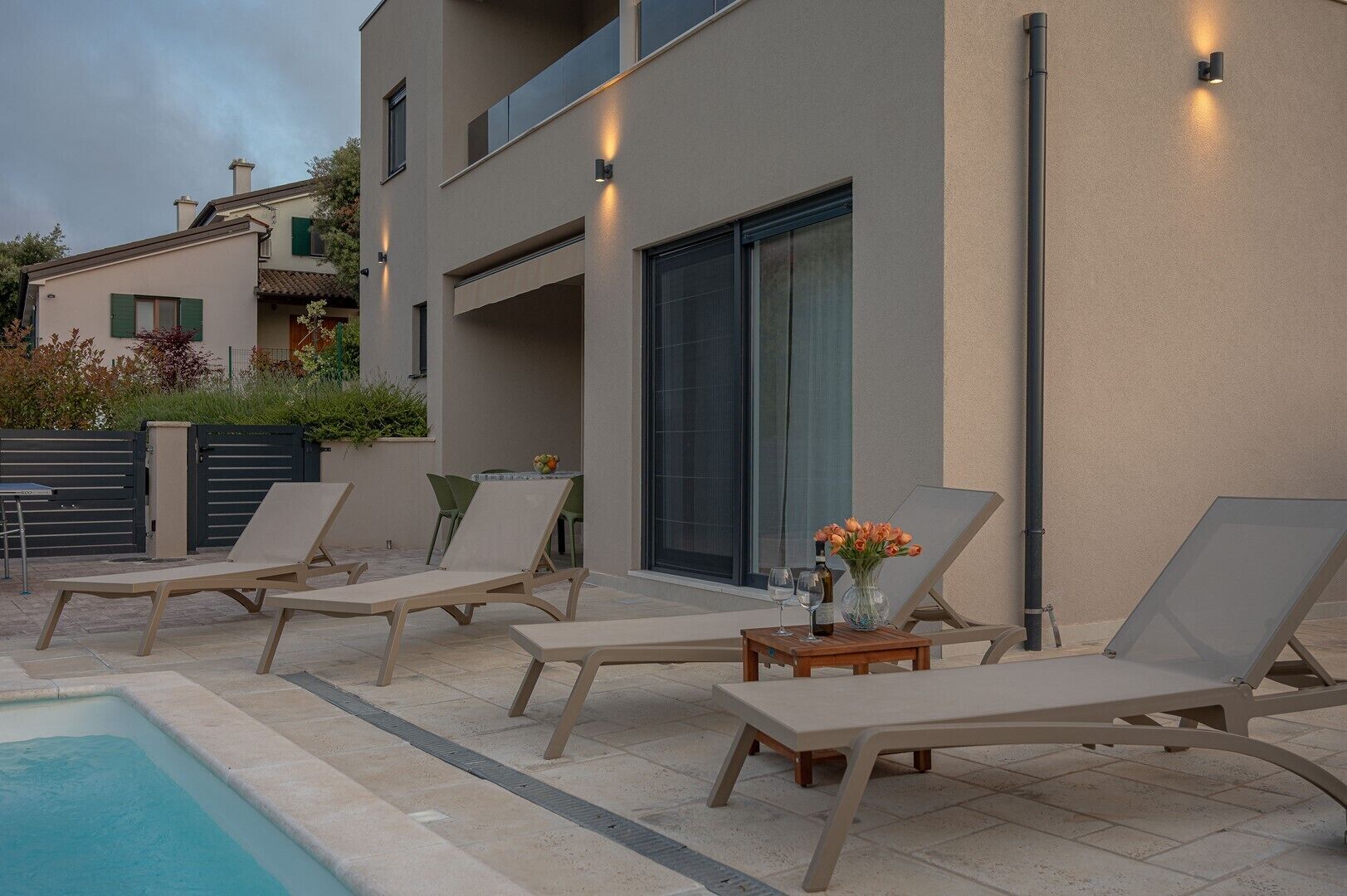 A modern outdoor pool area with lounge chairs and a table, surrounded by a building with large windows and outdoor lighting.