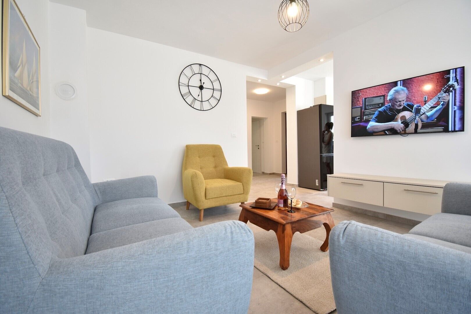 Een moderne woonkamer met twee grijze banken, een gele fauteuil, een houten salontafel met een fles en glazen, een wandklok en een televisie waarop een muzikant te zien is.