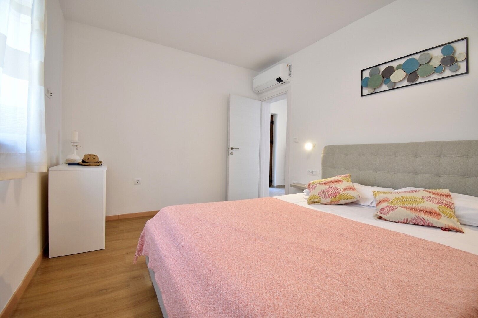 A bedroom with a bed, a bedside table, a wall-mounted air conditioner, and a door leading to another room.