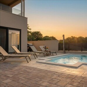 A modern outdoor pool area with lounge chairs and a table, surrounded by a low wall and a clear view of the sky.