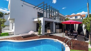 A modern outdoor pool area with a wooden deck, a hanging chair, and a red umbrella.