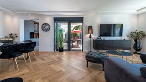 A modern living room with a wooden herringbone floor, a dining area with a black table and chairs, a large flat-screen TV, and a view of an outdoor patio with a red umbrella.