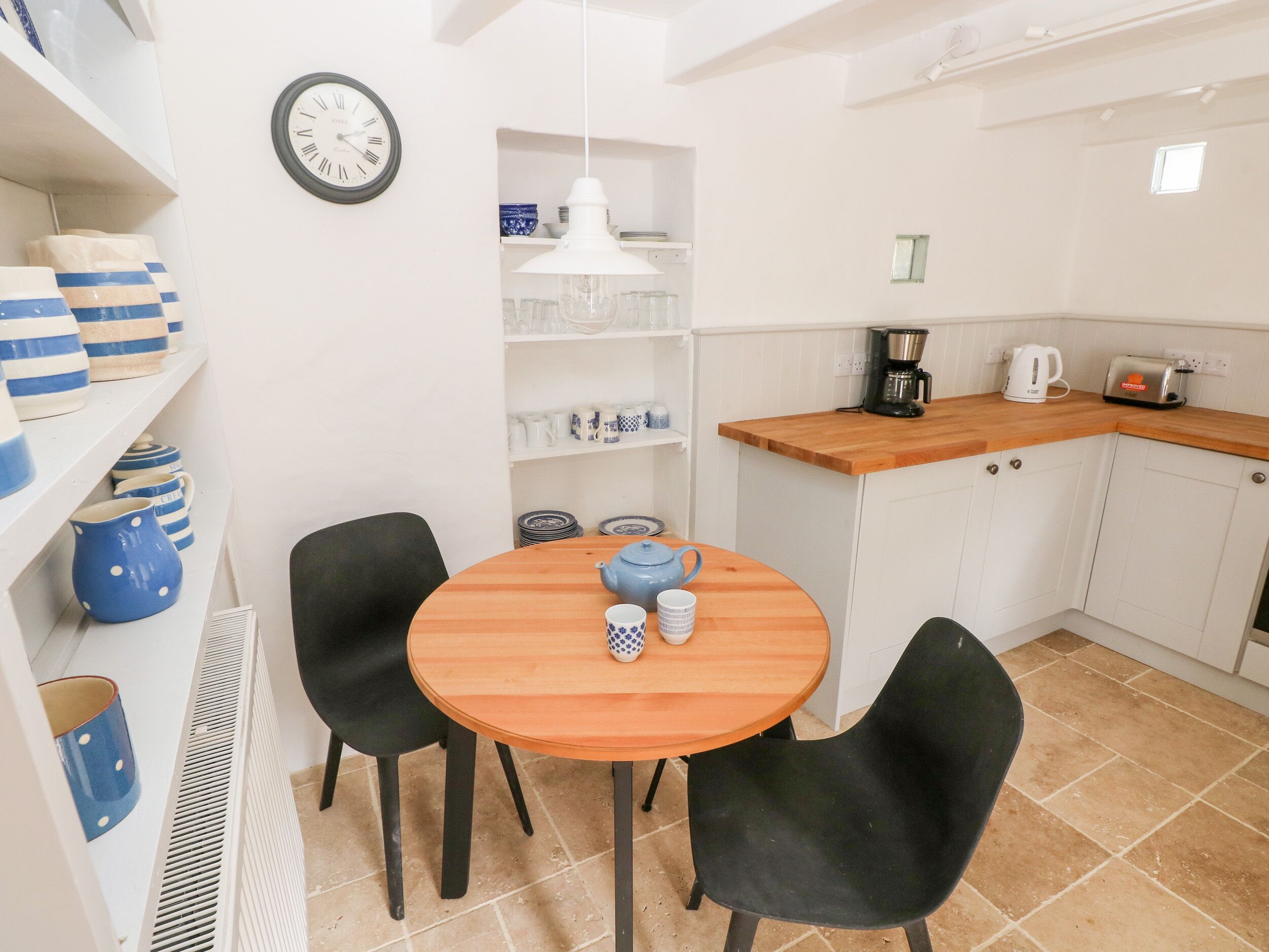 Una cocina compacta con una mesa redonda de madera, sillas negras, un reloj y diversos electrodomésticos.