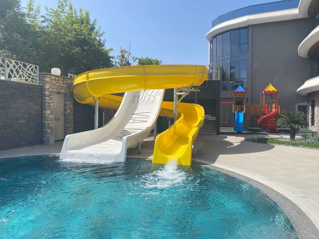 Una piscina con dos toboganes, uno blanco y otro amarillo, y un edificio con fachada curva al fondo.