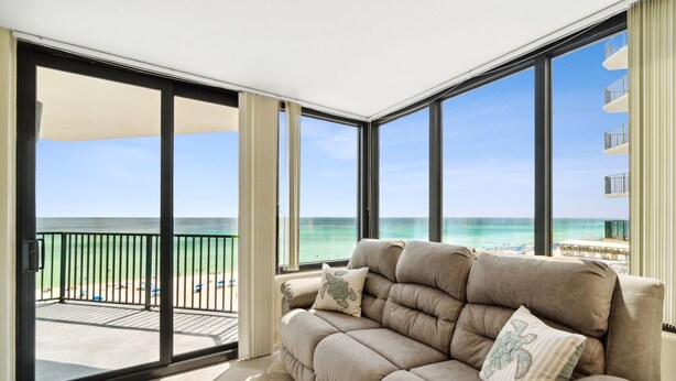 A balcony with a view of the ocean and beach, a beige sofa with decorative pillows, and a glass door leading to the balcony.