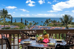 Et rundt metalbord med tallerkener, en ananas og en hibiscusblomst på en balkon med udsigt over havet.