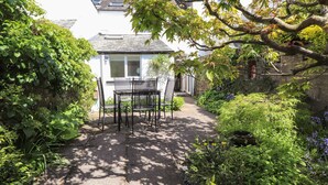 Ein Garten mit einer Terrasse, einem Tisch und Stühlen, umgeben von üppigem Grün und einem weißen Gebäude im Hintergrund.