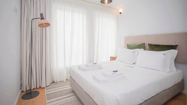 Ein ordentlich bezogenes Bett mit weißen Laken, eine Stehlampe und eine hängende Pendelleuchte in einem modernen Hotelzimmer.