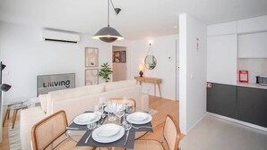 Ein modernes Wohnzimmer mit Essbereich, weißen Wänden, einer Couch, einem Esstisch gedeckt mit Tellern und Gläsern, einer Pendelleuchte und einer Küchenzeile mit Schränken und einem Feuerlöscher.