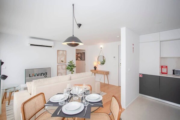 A modern living room with a dining area, white walls, a sofa, a dining table set with plates and glasses, a hanging light fixture, and a kitchen area with cabinets and a fire extinguisher.