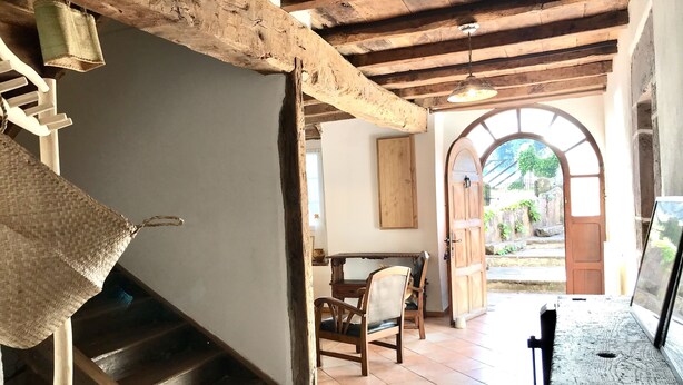 Ambiente rustico con travi a vista in legno, una scala, una porta in legno che dà sull’esterno e vista su un’area verde attraverso un arco.