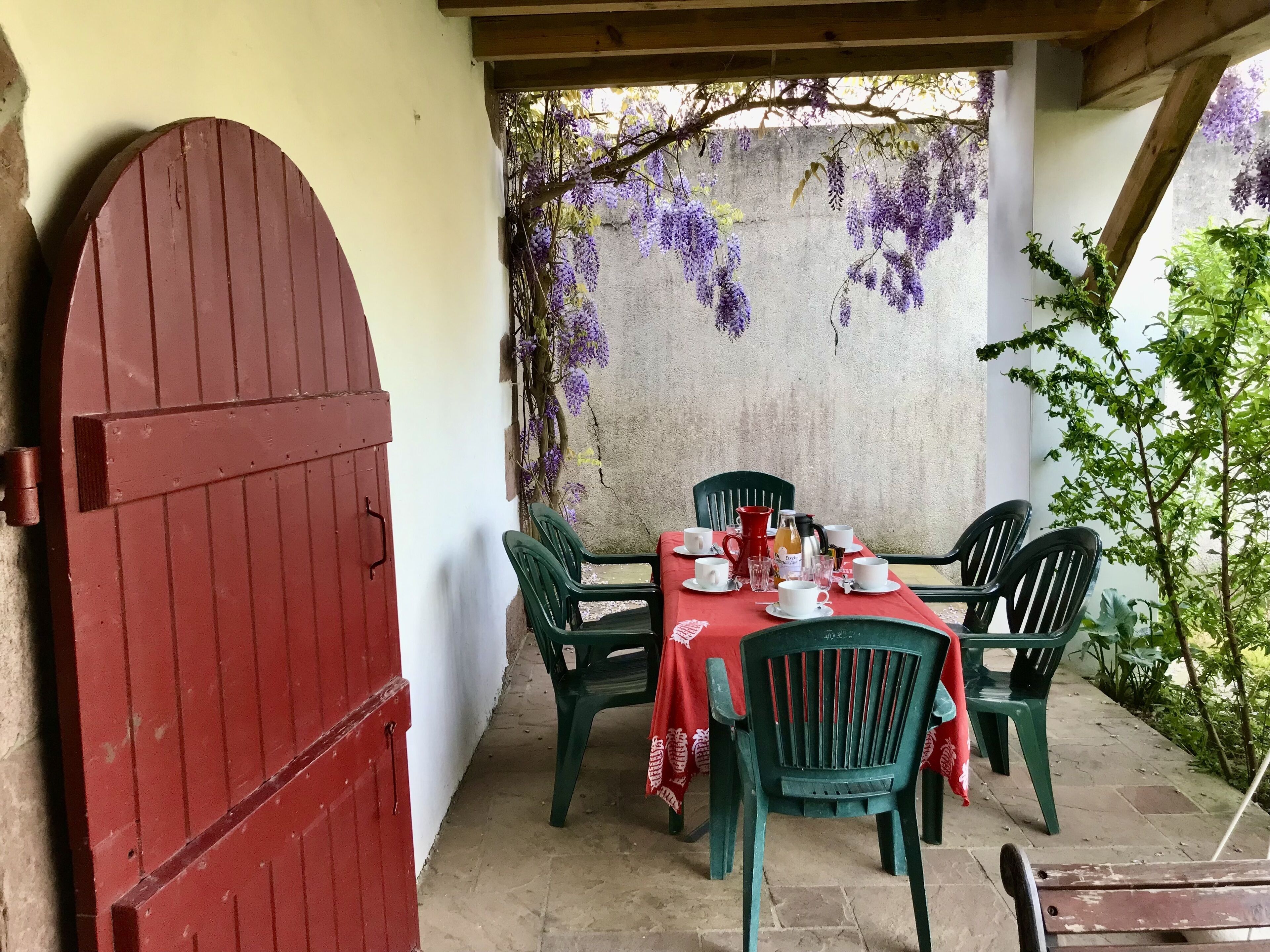 An outdoor patio with a table set for four, surrounded by green plastic chairs, a red door, and purple wisteria hanging from a wall.