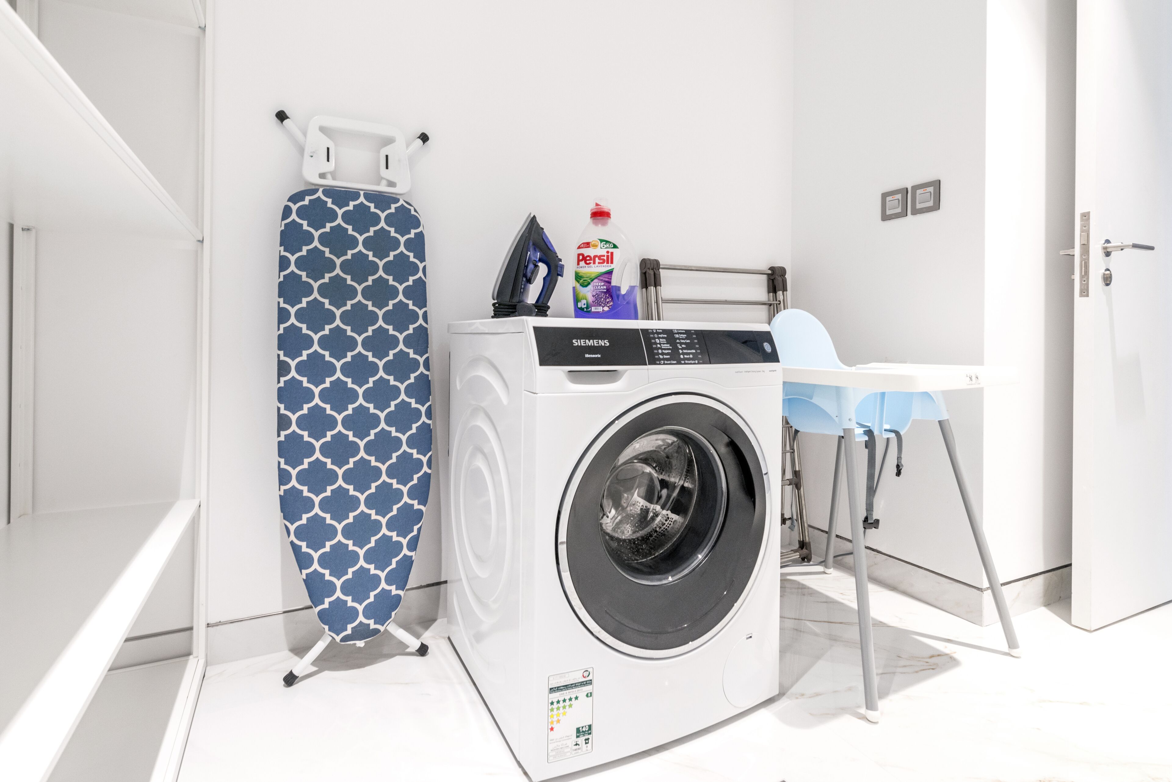 Een kleine wasruimte met een wasmachine, een strijkplank en een fles Persil wasmiddel.