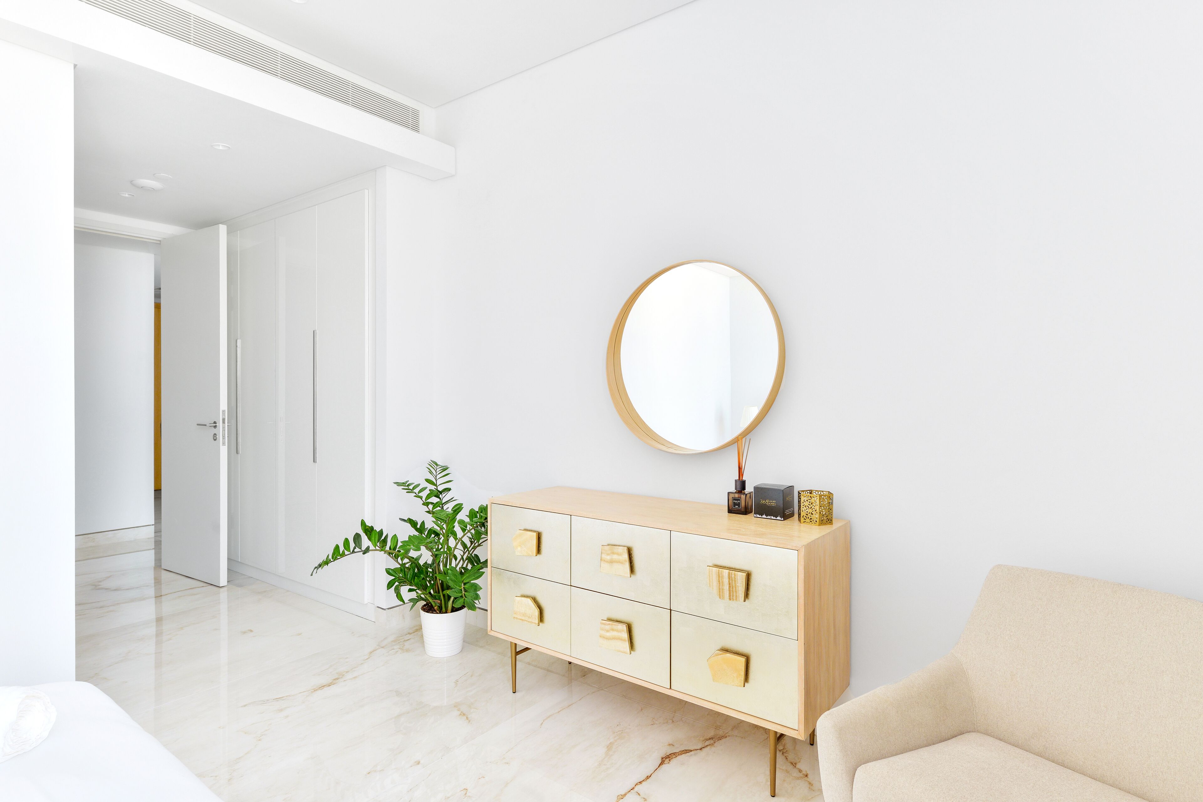 Una habitación moderna y minimalista con suelo de mármol, un sofá color beige, un mueble de madera con cajones, un espejo redondo y una planta en maceta.