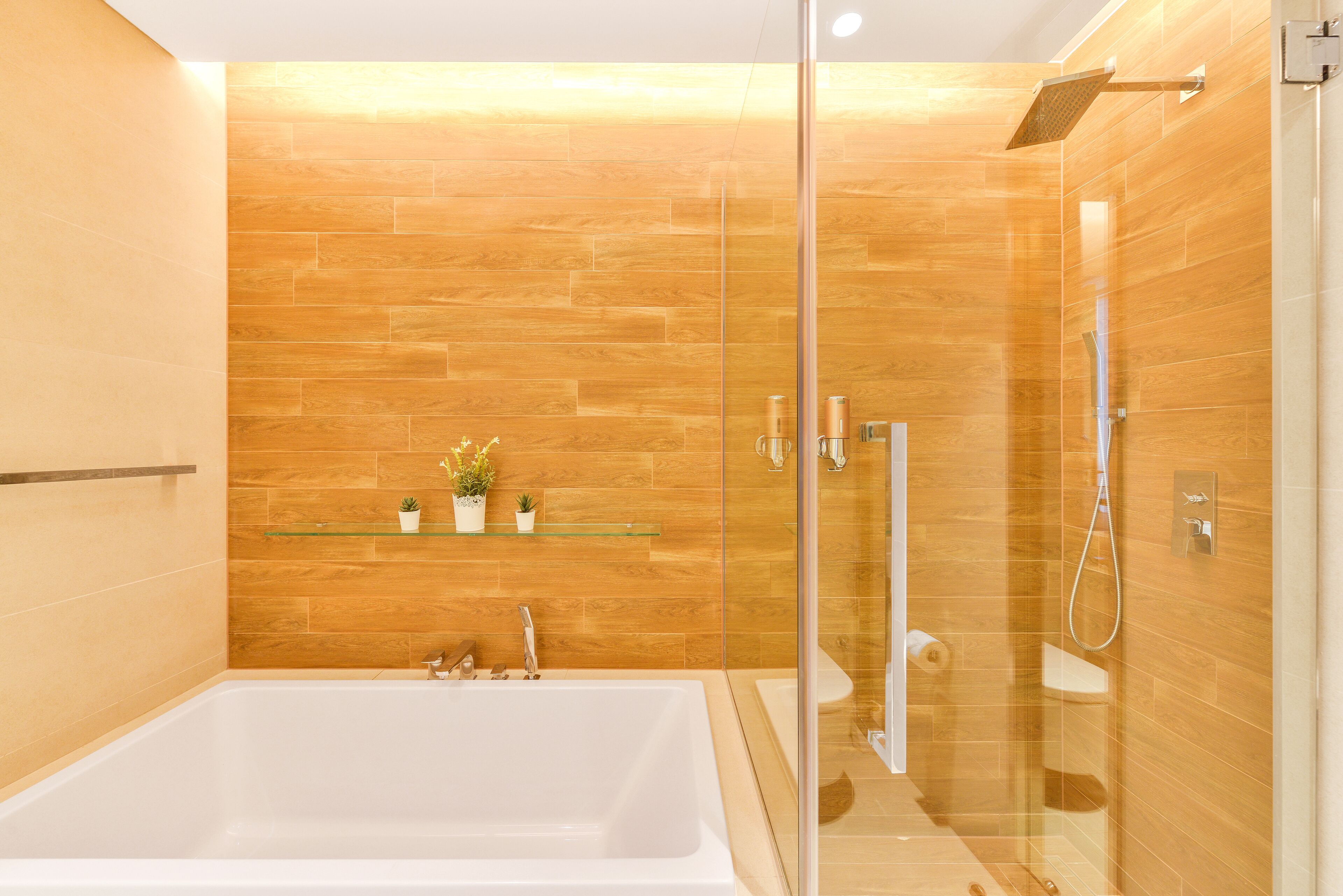 A modern bathroom with a glass-enclosed shower, a white bathtub, and wooden wall tiles.