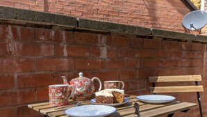 Une table en bois, dressée avec une théière, des tasses, un gâteau et des assiettes, le tout placé devant un mur de briques.
