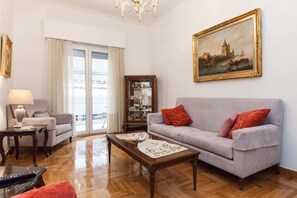 A living room with a grey sofa, red cushions, a wooden coffee table, a lamp, a cabinet with glass doors, and a painting of a cityscape. - Beautiful Retro Apt in Koukaki (Athens)
