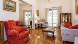 Amplia sala de estar con un sillón rojo, un sofá gris y una mesa de centro de madera. Se observa una lámpara de araña, un aparador con puertas de cristal y un cuadro en la pared.