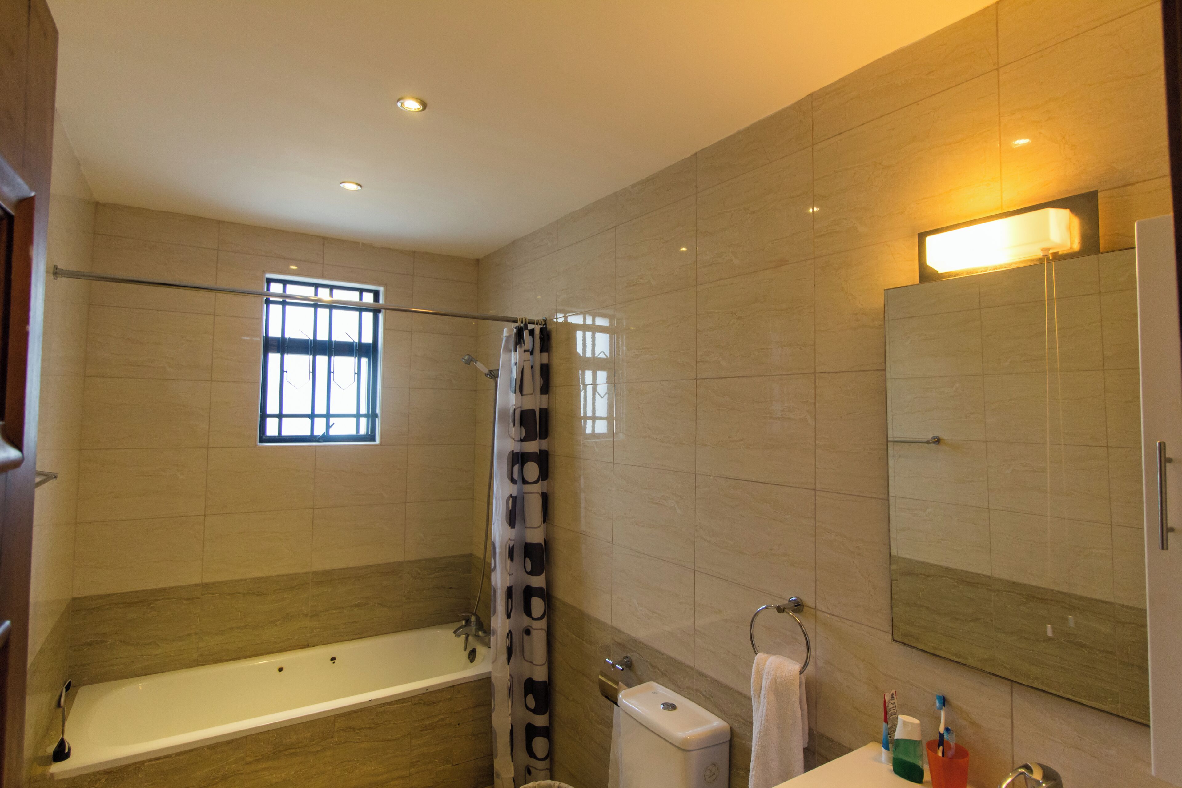 A modern bathroom with a bathtub, shower, and toilet. There is a window providing natural light and a mirror above the sink.