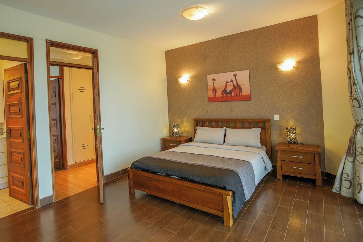 Quarto com cama de madeira, duas mesas de cabeceira com abajures, um quadro na parede e uma porta que dá para o banheiro.