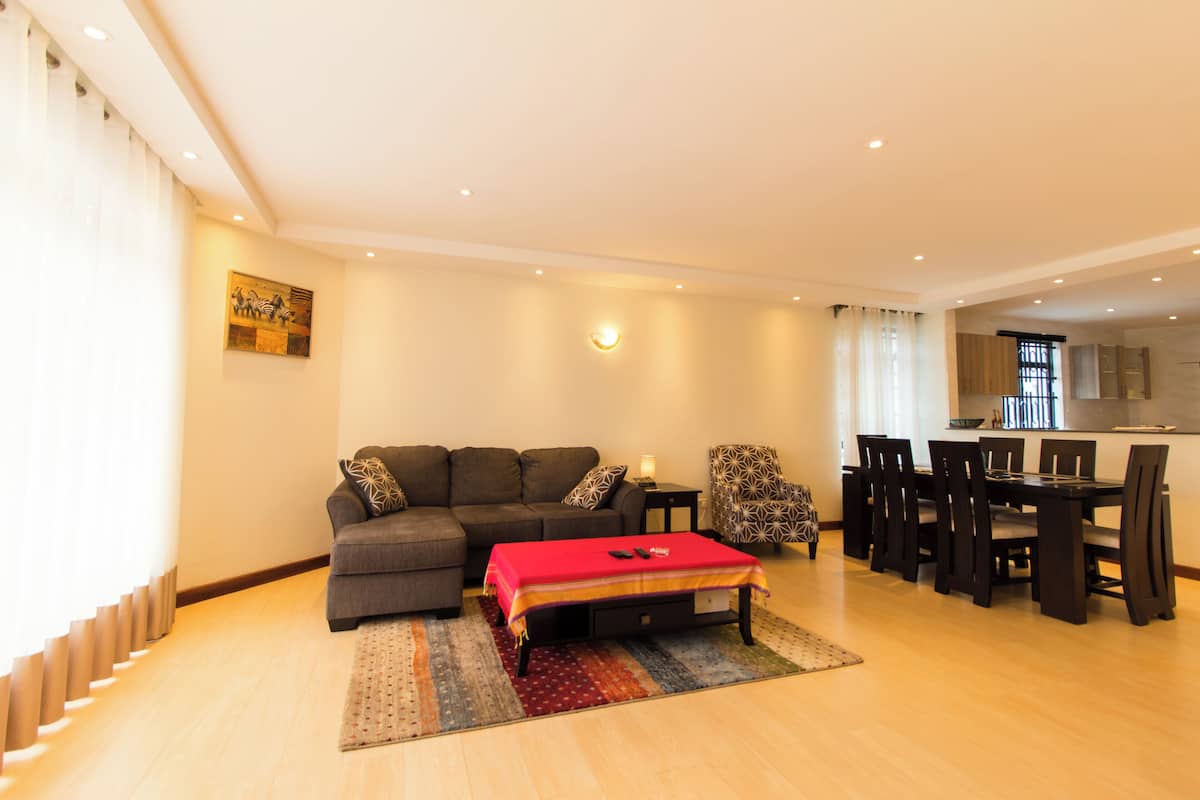 Uma sala de estar com sofá, mesa de jantar com cadeiras, uma mesinha de centro com abajur e um banquinho vermelho.