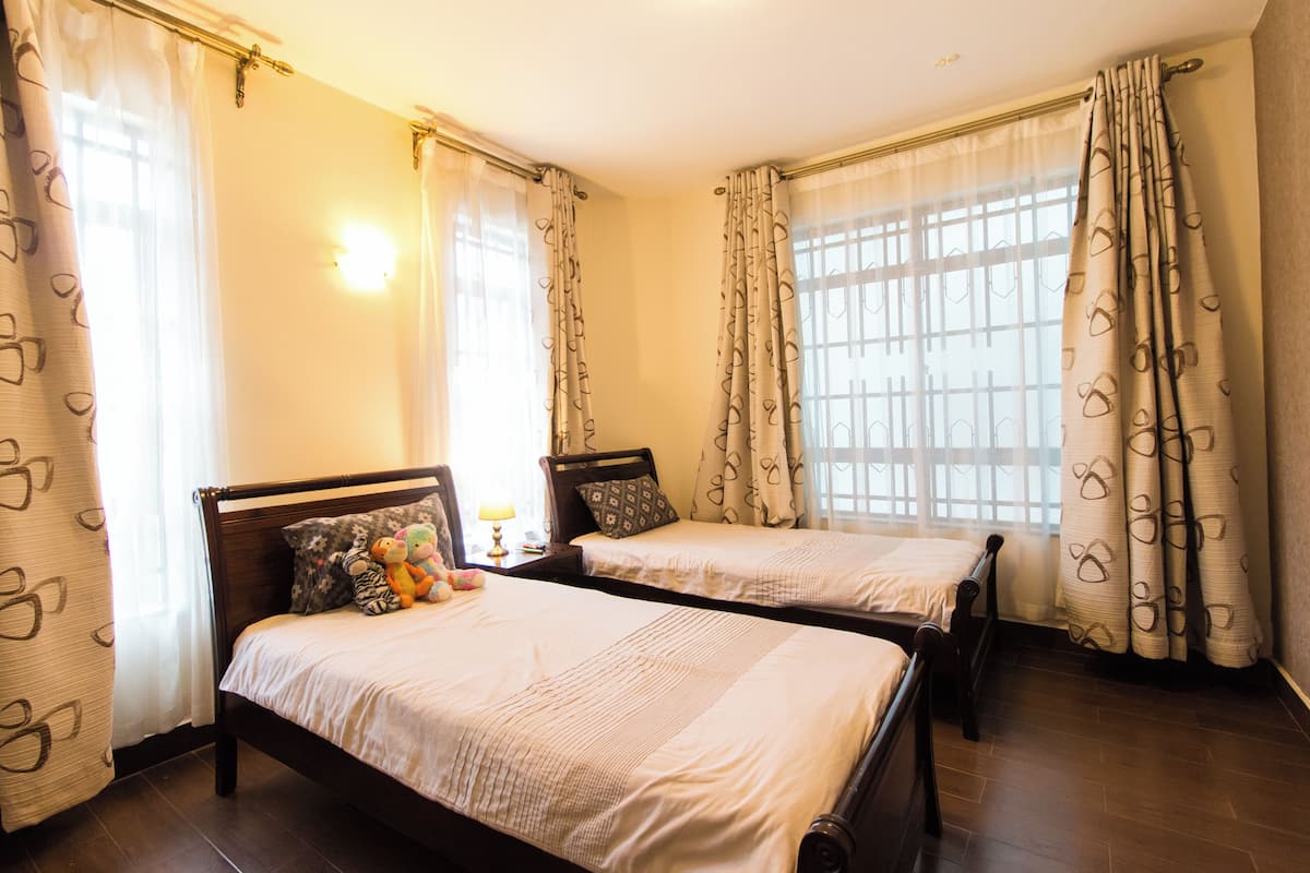 Quarto de hotel com duas camas de solteiro, uma mesa de cabeceira com abajur e uma janela com cortinas.