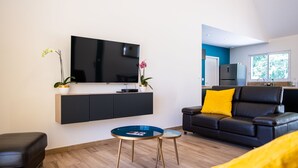 Un salon moderne avec un téléviseur à écran plat, un canapé noir agrémenté d’un coussin jaune, une petite table basse ronde et un pouf noir.
