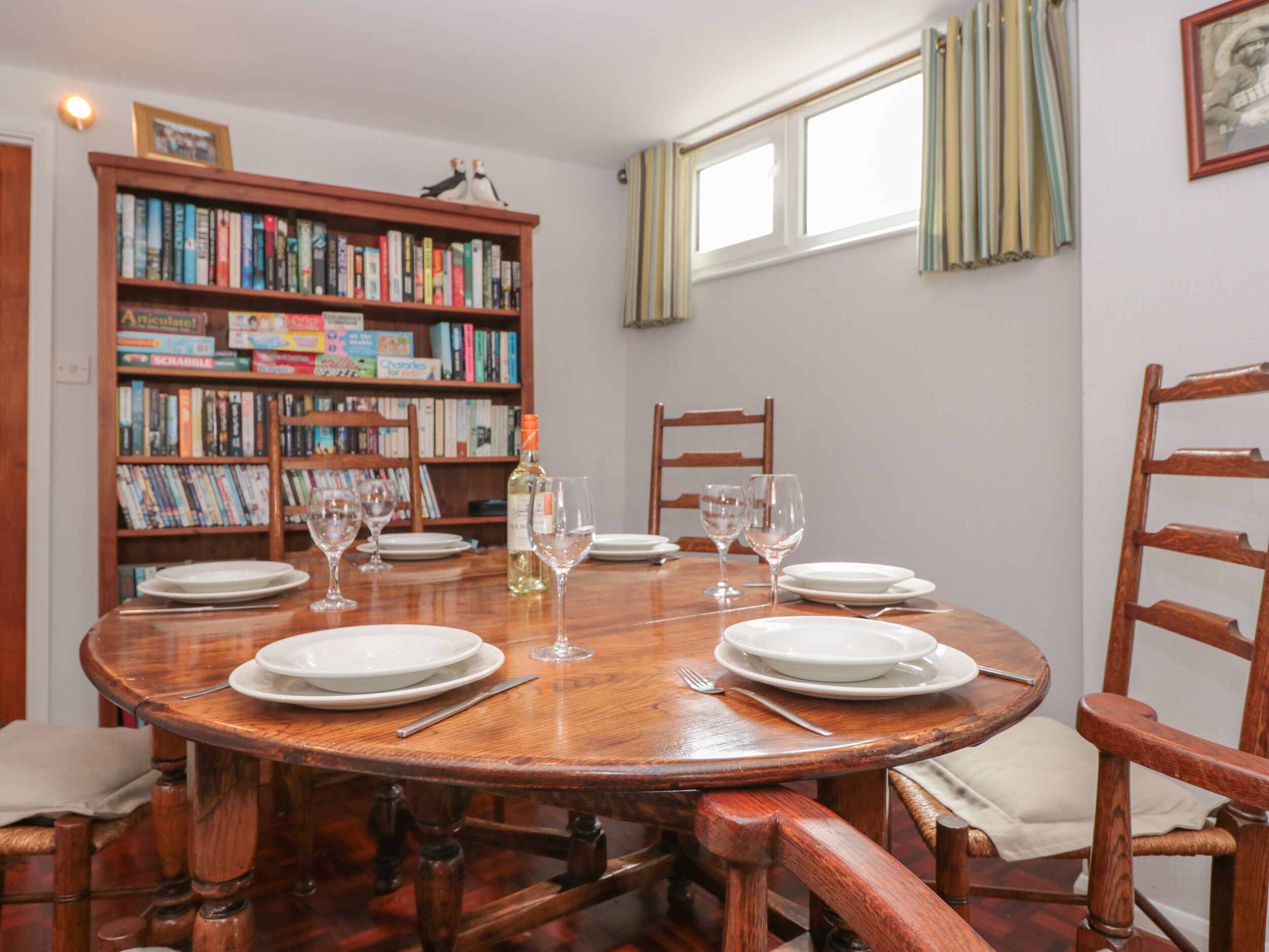 Una mesa de comedor redonda de madera con platos blancos y copas de vino, rodeada de sillas de madera. Un estante lleno de libros se ve al fondo.