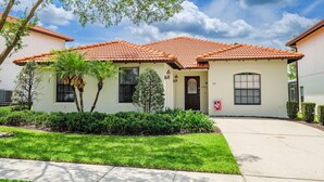 A two-story house with a red-tiled roof, a front door, and a small front yard with a palm tree and shrubs.