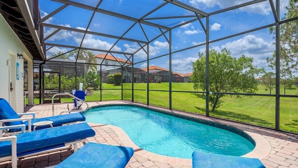 A swimming pool with blue lounge chairs, a covered area with a glass roof, and a view of a green landscape.
