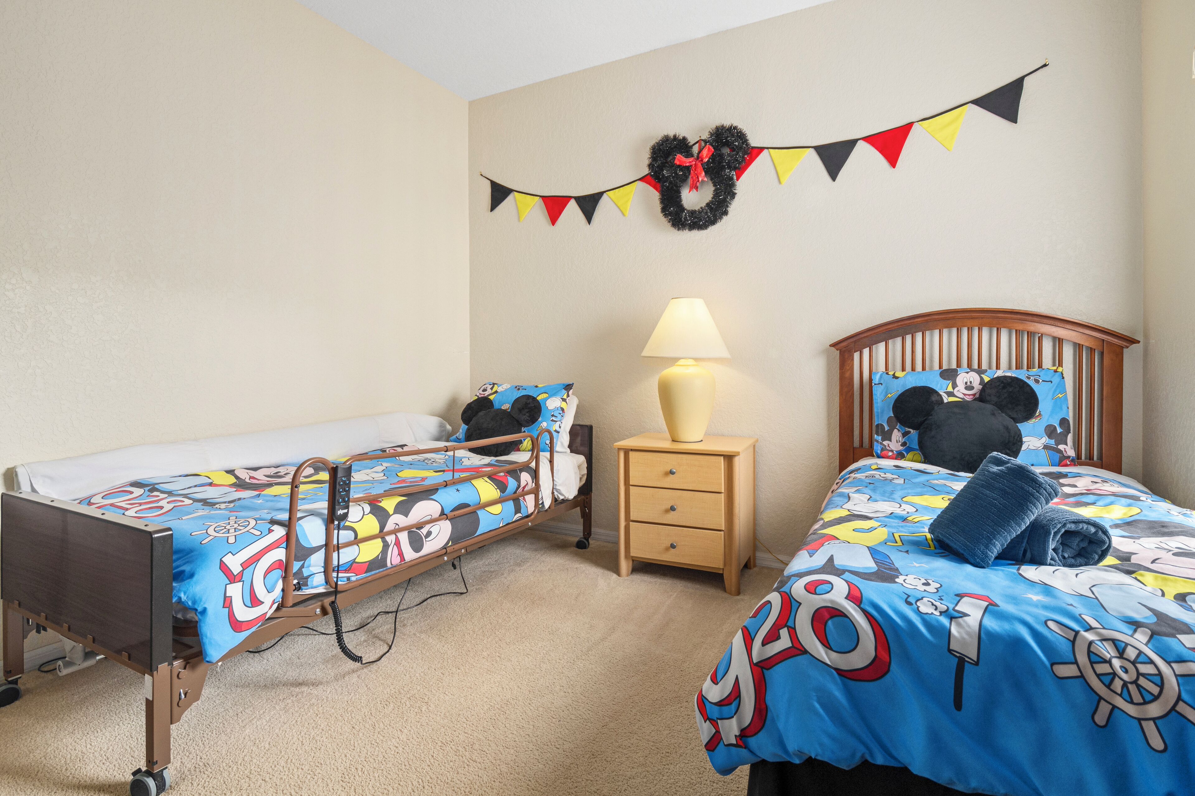 A hotel room with two beds, a nightstand with a lamp, a wall-mounted wreath, and Mickey Mouse-themed bedding.